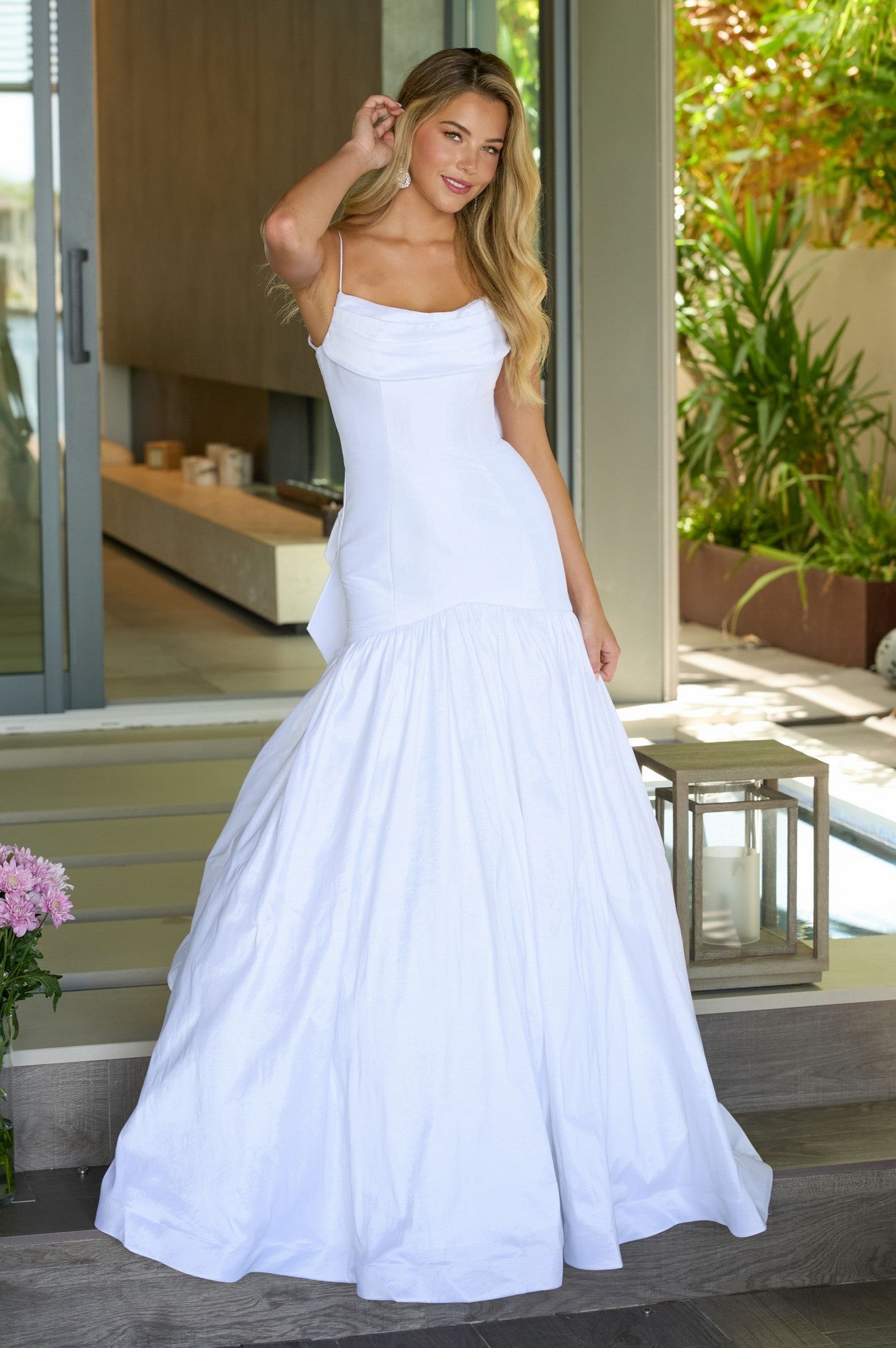 A woman in the Ava Presley 26422 Drop-Waist Mermaid Prom Gown stands indoors near a sliding door, smiling and holding her hair with one hand.